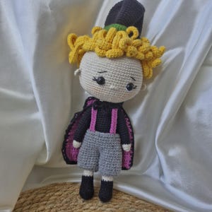 Könnte beinhalten: Eine gehäkelte Puppe mit gelben Haaren, einem schwarzen Hut und einem rosa-schwarzen Umhang. Die Puppe trägt eine graue Hose und schwarze Schuhe.