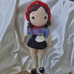 Könnte beinhalten: Eine gehäkelte Puppe mit braunen Haaren, die ein rotes Kapuzenkleid, ein blaues Oberteil, eine weiße Schürze und schwarze Schuhe trägt.