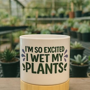 Pode incluir: Vaso de cerâmica branco com base de bambu. O vaso tem o texto "I'M SO EXCITED I WET MY PLANTS!" em verde e roxo. O fundo mostra uma estufa com plantas em vasos.
