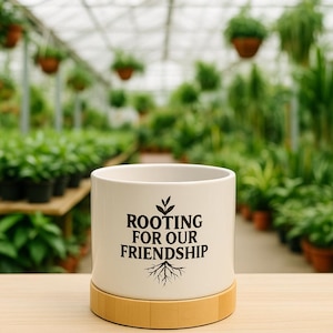 May include: A white ceramic planter with the text "ROOTING FOR OUR FRIENDSHIP" in black. The planter sits on a light brown wooden base. The background is a greenhouse filled with plants.