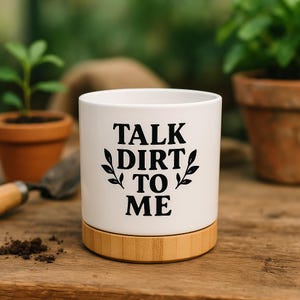 Puede incluir: Una maceta de cerámica blanca con base de bambú. La maceta tiene la frase "TALK DIRT TO ME" en letras negras con detalles de hojas. La maceta está sobre una superficie de madera, con otros elementos de jardinería en el fondo.