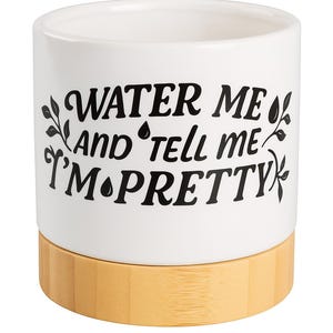 May include: White ceramic plant pot with a natural bamboo base. The pot features black text that reads "WATER ME AND TELL ME I'M PRETTY" with decorative leaf and water drop accents. A decorative container for plants.