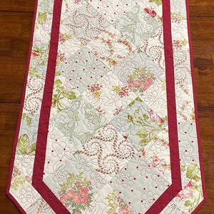 May include: A quilted table runner with a floral pattern in shades of pink, green, and white. The runner has a burgundy border with a double-stitched edge.