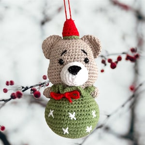Puede incluir: Un adorno de oso de peluche tejido a crochet con un cuerpo a rayas verde y blanco, un lazo rojo y un gorro de Papá Noel rojo. El adorno está colgado de una rama con bayas rojas.
