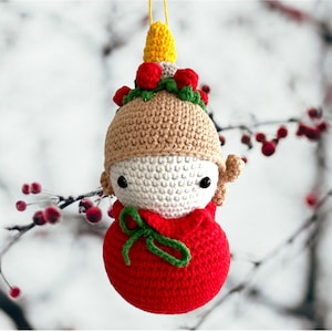 May include: A crocheted Christmas ornament shaped like a person wearing a brown hat with a yellow and red top, a white face, and a red dress with a green bow.