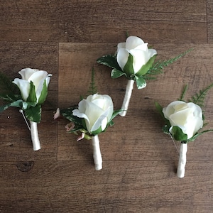 May include: Four white rose buttonholes with green leaves and white ribbon. The buttonholes are arranged on a brown wooden surface.