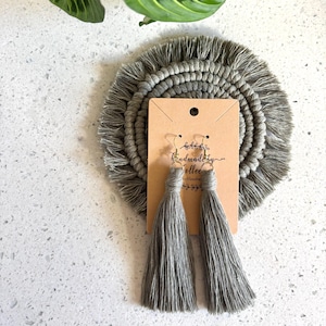 Puede incluir: Un par de pendientes de borla gris con ganchos dorados. Los pendientes se muestran en una tarjeta que dice "Colección hecha a mano" y están sobre un posavasos de macramé gris.