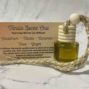 Puede incluir: Una pequeña botella de vidrio con una tapa de madera y una cuerda dorada y blanca colgando de ella. La botella está llena de un líquido transparente y tiene una etiqueta que dice "Vanilla Spiced Chai Rearview Mirror Car Diffuser Cardamom - Vanilla - Cinnamon - Clove - Ginger". Para difundir: Voltea la botella para permitir que el aceite se filtre de la tapa de madera, luego cuélgala del espejo retrovisor y ata la cuerda. Voltea la botella cuando el aroma necesite ser refrescado. ¡Repite tan a menudo como sea necesario! Usa la botella, retira la tapa de plástico de la botella