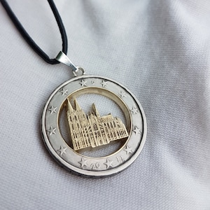 Könnte beinhalten: Eine Halskette aus Silber und Gold mit einem Anhänger, der eine detaillierte Ausschnitt eines Kathedralengebäudes zeigt. Der Anhänger ist von einem Kreis aus Sternen und der Inschrift "20" umgeben.