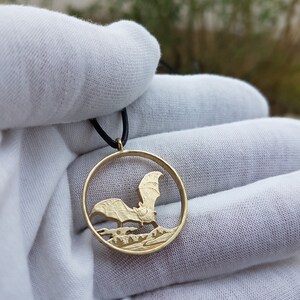 May include: A gold-colored coin necklace with a bat cut-out design. The bat is flying over a forest scene. The necklace is on a black cord.