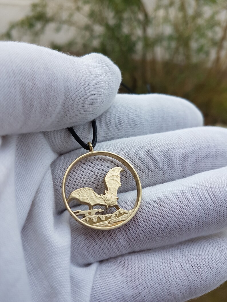 May include: A gold-colored coin necklace with a bat cut-out design. The bat is flying over a forest scene. The necklace is on a black cord.