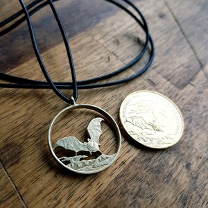 May include: A silver bat pendant necklace with a black cord. The pendant is a cut-out of a bat flying over a forest scene. The back of the pendant is a gold coin with the inscription "PODKOWIANY PARK NARODOWY" and a bat flying over a forest scene.