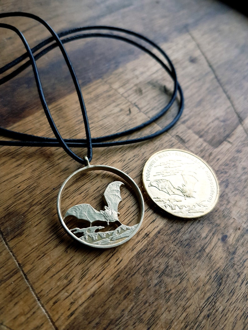 May include: A silver bat pendant necklace with a black cord. The pendant is a cut-out of a bat flying over a forest scene. The back of the pendant is a gold coin with the inscription "PODKOWIANY PARK NARODOWY" and a bat flying over a forest scene.