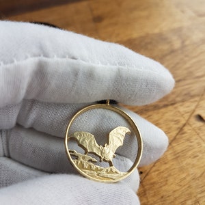 May include: A gold-colored coin pendant featuring a detailed cutout of a bat in flight. The bat is silhouetted against a landscape with trees and a moon.