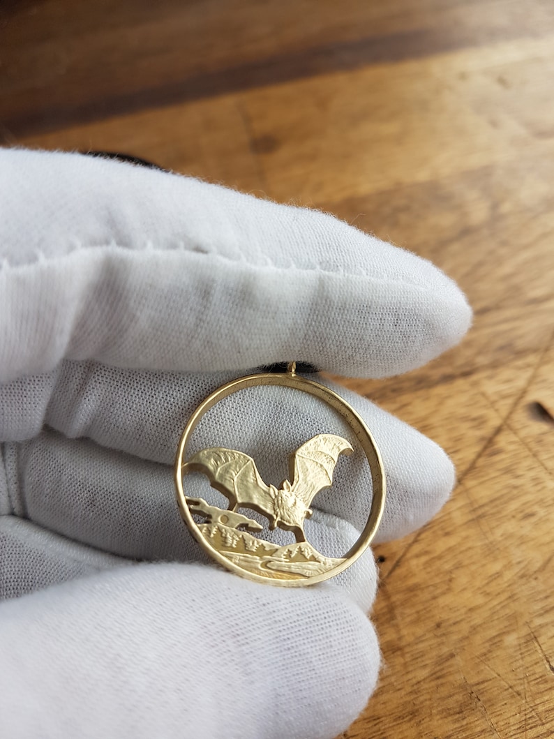 May include: A gold-colored coin pendant featuring a detailed cutout of a bat in flight. The bat is silhouetted against a landscape with trees and a moon.
