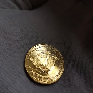 May include: A gold coin with a bat in flight, surrounded by clouds. The coin has the inscription "PODKOWIEC MAŁY RHINOLOPHUS HIPPOSIDEROS" around the edge.