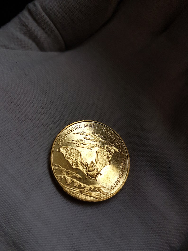 May include: A gold coin with a bat in flight, surrounded by clouds. The coin has the inscription "PODKOWIEC MAŁY RHINOLOPHUS HIPPOSIDEROS" around the edge.