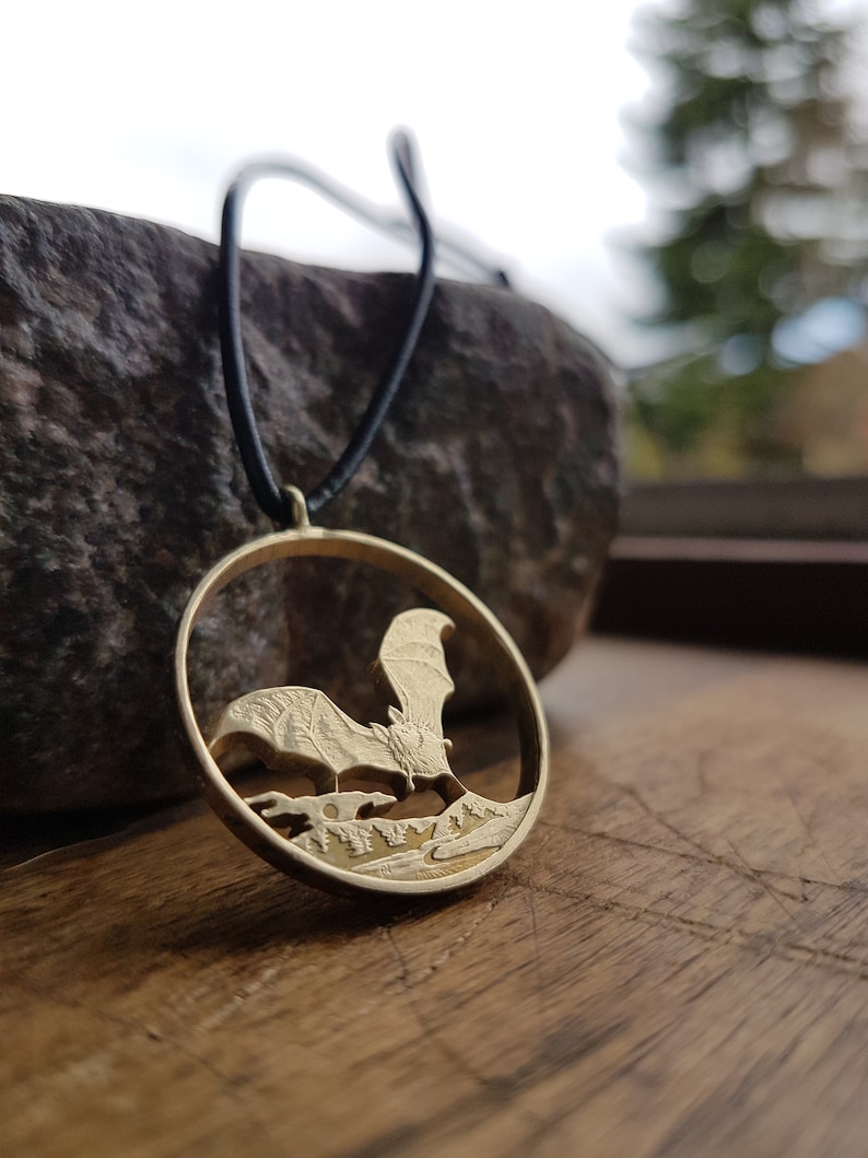 May include: A gold-toned necklace with a round pendant featuring a detailed silhouette of a bat flying over a forest scene.
