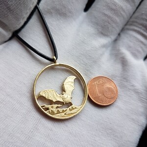 May include: A gold-colored pendant necklace featuring a detailed cutout of a bat in flight. The pendant is circular and is about the size of a 1-cent Euro coin.
