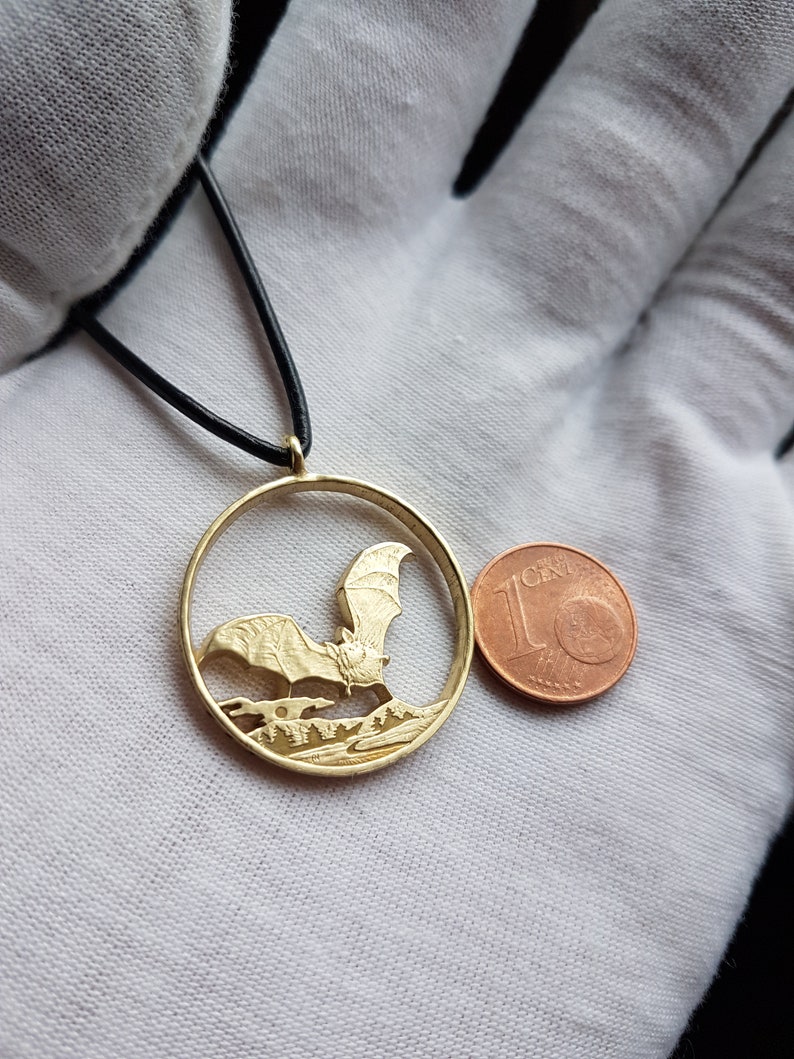 May include: A gold-colored pendant necklace featuring a detailed cutout of a bat in flight. The pendant is circular and is about the size of a 1-cent Euro coin.