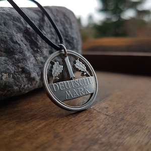Könnte beinhalten: Eine silberne Anhänger-Halskette mit einem ausgeschnittenen Design. Der Anhänger trägt die Aufschrift "DEUTSCHE MARK" und Eichenblattdetails. Er hängt an einer schwarzen Kordel. Die Halskette wird auf einer Holzoberfläche präsentiert.