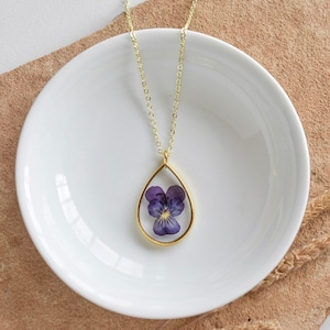 May include: A gold chain necklace with a teardrop-shaped pendant featuring a pressed purple flower.