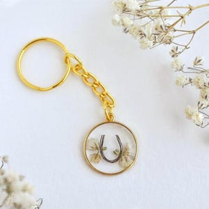 May include: A gold-coloured keyring with a round pendant featuring dried flowers and a horseshoe charm. The keyring has a gold ring and chain. White flowers are in the background.