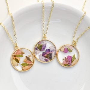 May include: Three gold necklaces with round pendants featuring pressed flowers encased in clear resin. The flowers are in shades of pink, purple, and white.