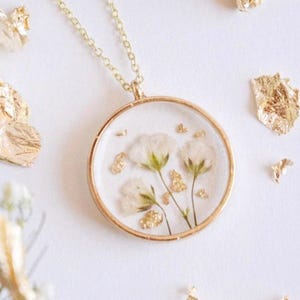 May include: A gold chain necklace with a round pendant featuring pressed white flowers and gold leaf accents.