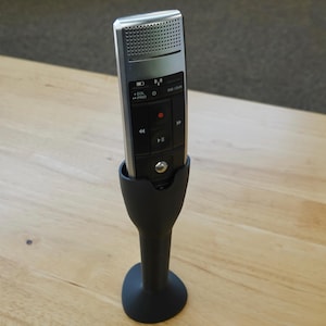 May include: A silver and black digital voice recorder is held in a black stand. The recorder has a microphone, buttons, and a small screen. The stand is shaped like a goblet and is on a light brown wooden surface.