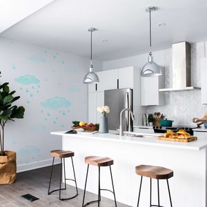 May include: A white kitchen island with a white countertop and three wooden bar stools. The island has a stainless steel sink and faucet. The kitchen has white cabinets and a stainless steel range hood. The wall behind the island has a light blue and white watercolor design of clouds and raindrops.