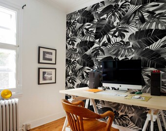 Op de afbeelding: Een thuiskantoorwerkplek met een wit bureau en een houten stoel. De muur achter het bureau is bedekt met zwart-witte tropische bladbehang. Een computermonitor staat op het bureau, samen met een toetsenbord en andere kantoorbenodigdheden.