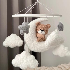 Puede incluir: Móvil para bebé hecho a mano con un osito de peluche marrón metido en una luna creciente blanca y esponjosa. El móvil incluye estrellas grises y acentos de nubes blancas, suspendidos de un marco circular blanco. La decoración de la guardería es suave y caprichosa.