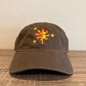 May include: A brown baseball cap with an embroidered yellow sun with red and orange rays, a yellow and orange cross, and five yellow stars.