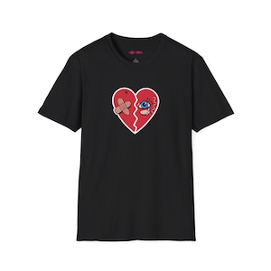 May include: Black t-shirt with a graphic of a red, broken heart with a bandage and an eye. The heart has a white outline and is stitched together with red thread. The shirt has a crew neck and short sleeves.