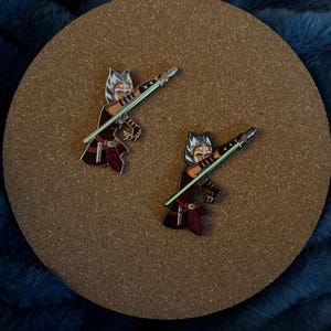 May include: Two enamel pins featuring a cartoon character with a lightsaber. The character is depicted in a dynamic pose, with a green lightsaber. The pins have a maroon and black color scheme, set against a cork background.