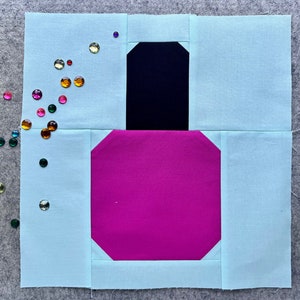 Op de afbeelding: Een lichtblauw vierkant van stof met een roze en zwart stoffen ontwerp in het midden. Het ontwerp lijkt op een nagellak flesje. Er zijn kleurrijke strass steentjes rondom het vierkant van stof gestrooid.