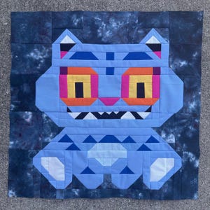 May include: A handmade quilt featuring a geometric design of a blue cat-like creature. The cat has orange and yellow eyes, pink accents, and a white-toothed grin. The background is a dark blue tie-dye pattern.