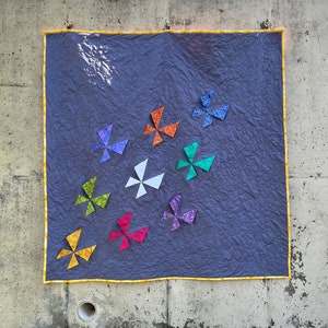 May include: A quilt with a dark grey background and a yellow border. The quilt features nine colourful pinwheels in various shades of red, orange, yellow, green, blue, and purple.