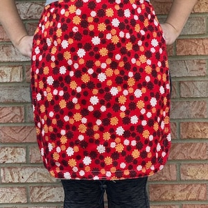 Puede incluir: Un delantal rojo con un estampado floral blanco y naranja. El delantal tiene una cintura a rayas blancas y negras.