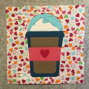 May include: A paper coffee cup with a pink heart on the side, surrounded by a white background with a pattern of colorful hearts.