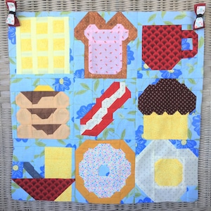 Puede incluir: Un fondo azul con una colcha de retazos de varios alimentos para el desayuno. La colcha incluye un waffle, tostadas, una taza de café, una pila de panqueques, tocino, un cupcake, una dona y un huevo.
