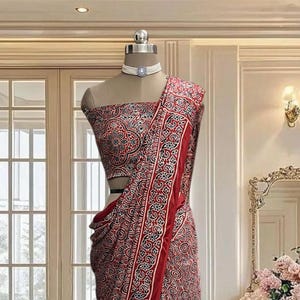 May include: A saree featuring a deep red base with an intricate repeating pattern in black and white. The garment includes a matching sleeveless blouse with a straight neckline and a white choker necklace with a central blue stone accent.