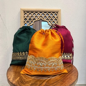 May include: Three drawstring pouches in dark green, bright orange, and deep magenta. Each pouch features a textured fabric finish and a decorative gold-toned embroidered trim along the bottom edge with intricate floral and sequin detailing.