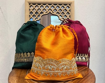 Könnte beinhalten: Drei Kordelzugbeutel in Dunkelgrün, leuchtendem Orange und tiefem Magenta. Jeder Beutel hat eine strukturierte Stoffoberfläche und eine dekorative, goldfarbene Stickborte am unteren Rand mit filigranen Blumen- und Paillettenverzierungen.