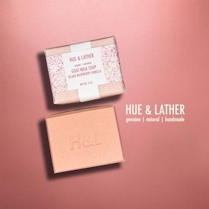 May include: Two bars of goat milk soap on a pink background. One bar is wrapped with a label that reads "HUE & LATHER GOAT MILK SOAP BLACK RASPBERRY VANILLA NET WT. 6 OZ." The other bar is peach-colored with "H&L" embossed on it.
