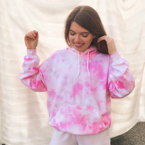 Può includere: Una felpa con cappuccio rosa e bianca tie-dye con tasca frontale e coulisse bianca. La felpa ha maniche lunghe e cappuccio. Il motivo tie-dye presenta sfumature di rosa e bianco.