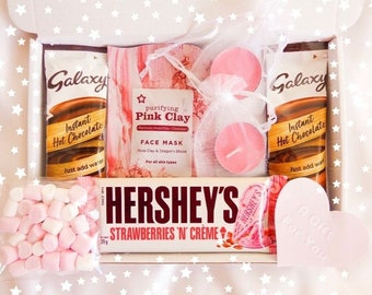 May include: A pink and white gift box filled with a variety of items, including a pink clay face mask, two pink tea lights, a package of instant hot chocolate, a chocolate bar, marshmallows, and a pink heart-shaped soap. The box is decorated with white stars.