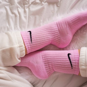 May include: A pair of pink athletic socks with the Nike swoosh logo in black. The socks are ribbed and worn with white sweatpants. The image is taken from a low angle, with a soft, white background.