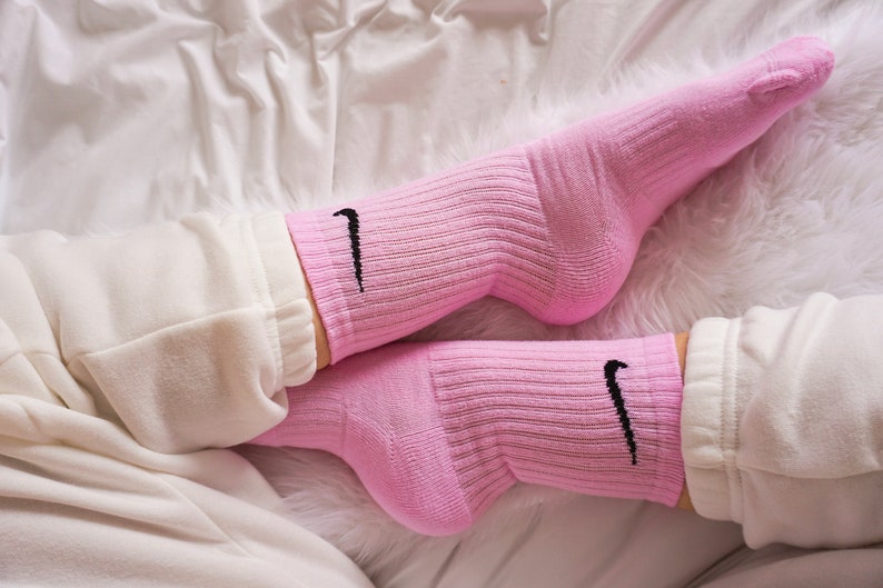 May include: A pair of pink athletic socks with the Nike swoosh logo in black. The socks are ribbed and worn with white sweatpants. The image is taken from a low angle, with a soft, white background.